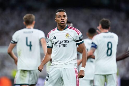 كيليان مبابي بقميص ريال مدريد يحمل الكرة الذهبية، مع شعار ريال مدريد في الخلفية.
